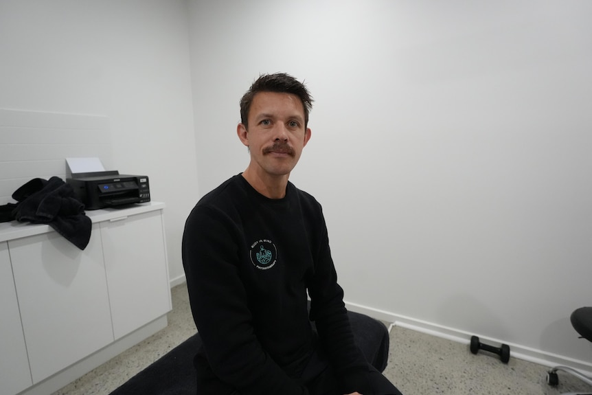 A headshot of a man with a moustache sitting on a treatment table in a physiotherapy office.