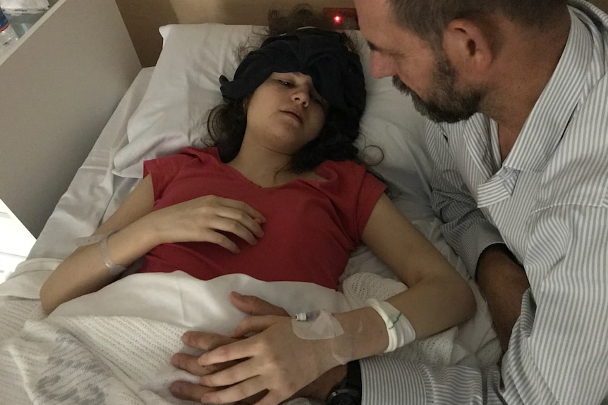 A young girl lies in bed, with her dad looking over her.