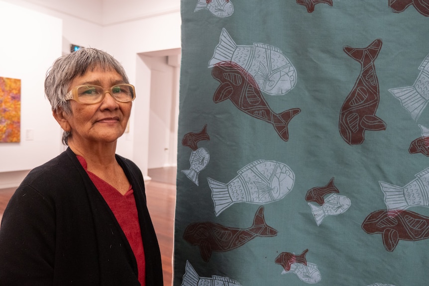 Woman with short hair and black jacket stands next to printed silk panel with block printed fish design