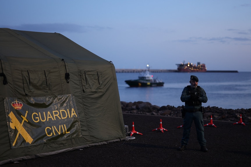 A member of the Spanish Civil Guard stands near some traffic cones in front of the port, next to a tent. 