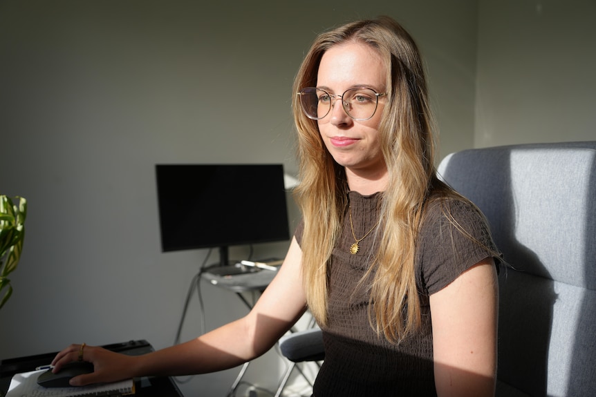A woman with long hair and glasses sits and looks at a computer.