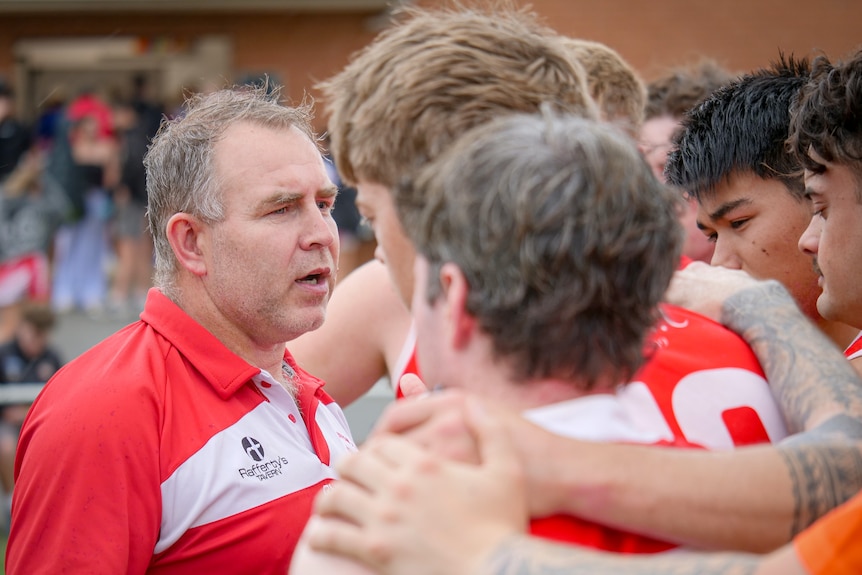 A coach with short greying hair talks to footballers with their arms around each other