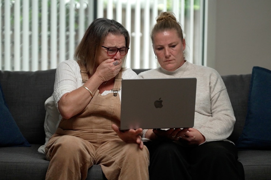 Two women sit on a couch looking at a laptop. One holds her hand to her mouth in horror, crying. 