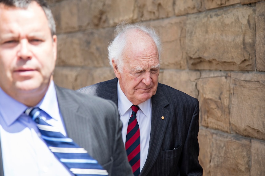 An older man in a suit walking in front of a stone wall