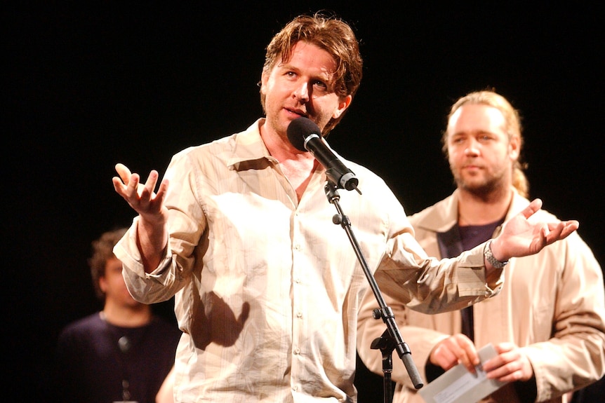 John Polson gestures as he stands at a microphone. Russell Crowe, with blonde ponytail, stands behind him.