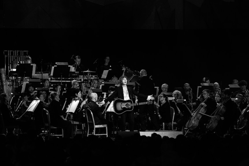 Black & White photo of suited male musician with acoustic guitar in front of a large symphony orchestra on stage