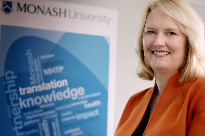 A smiling blonde woman in front of a Monash university promotional banner.