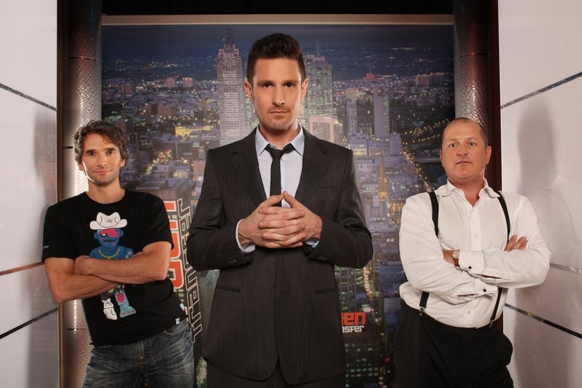 Wil, Todd and Russel stand in a hallway before an image of a city skyline with Wil in front steepling his hands.