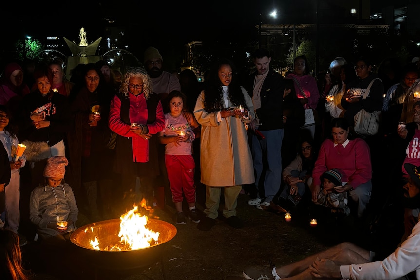 Lots of people at a vigil, with a fire pit in the centre.