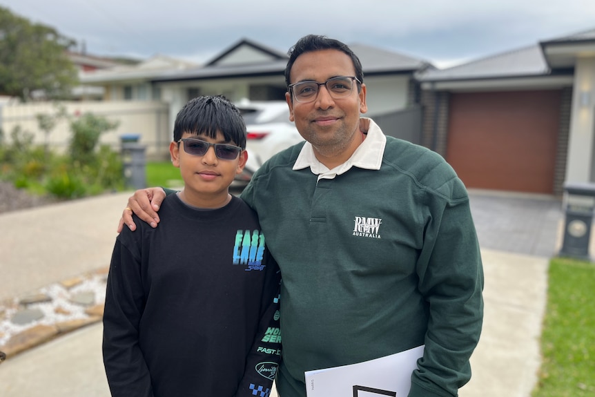 A man with his arm on the shoulder of a boy standing in front of a house and car