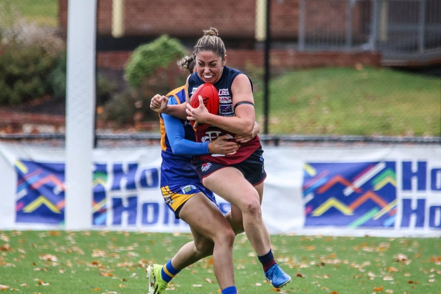 A female Aussie Rules player holding a red ball is tackled.