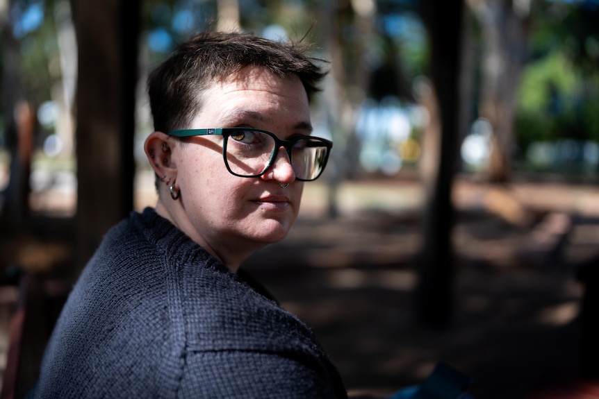 Le with glasses sits outdoors in a park, turns to look at the camera, serious, background blurred.