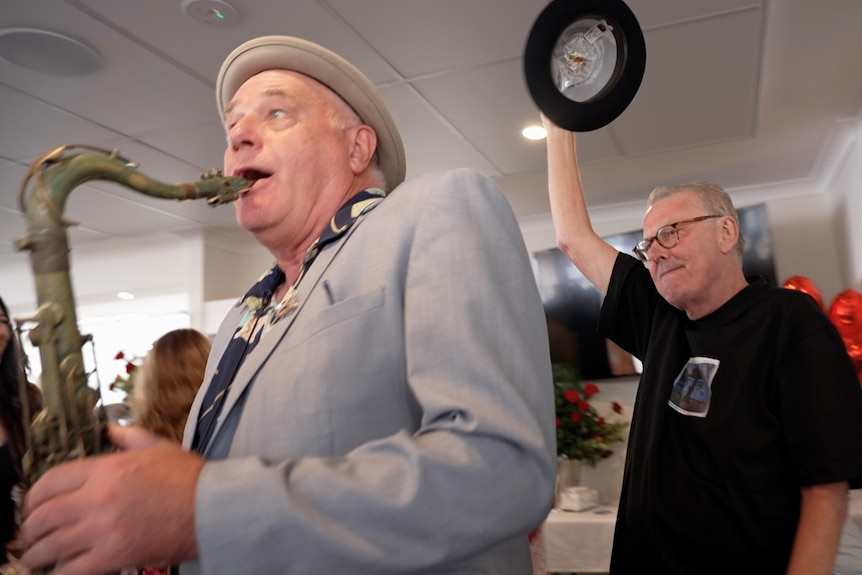 A man in black T-shirt holds a bowler hat high in the air as he walks into a room. A man in grey suit plays saxophone ahead