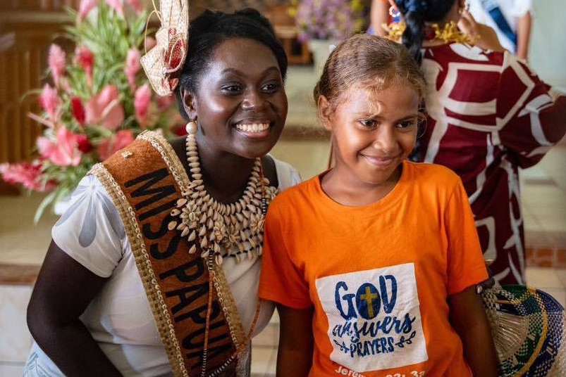 A young woman wearing a 'Miss Papua New Guinea' sash stands next to a smiling child posing for a photo