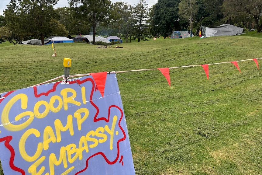 A purple sign in a grassy area reads in yellow writing: Goori camp embassy, with a red outline.