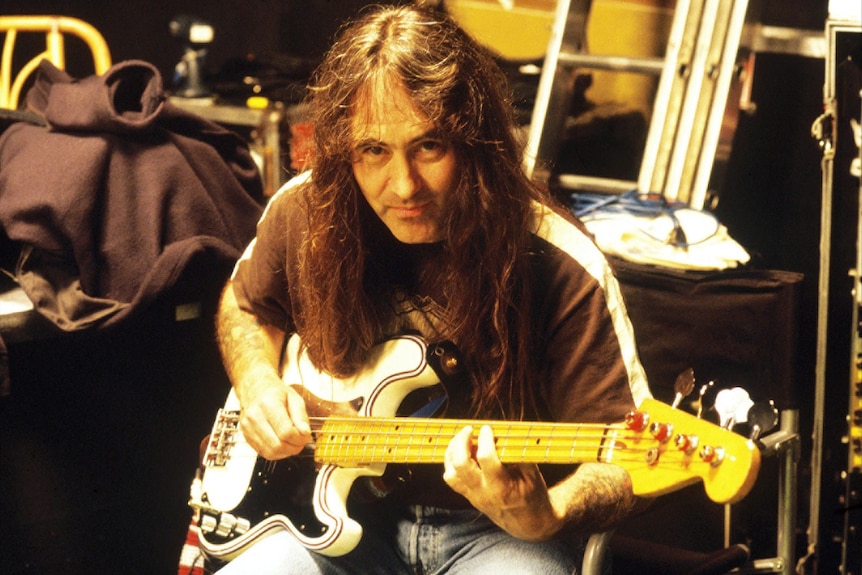 Person with long hair playing an electric bass guitar in a dimly lit backstage or studio setting.
