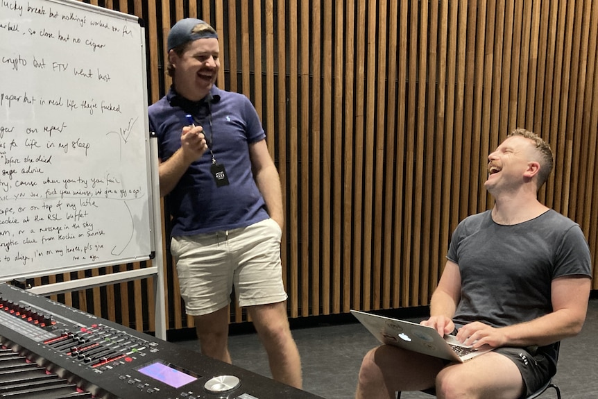 man stands next to a white board, looking at man who is sitting down with a laptop.