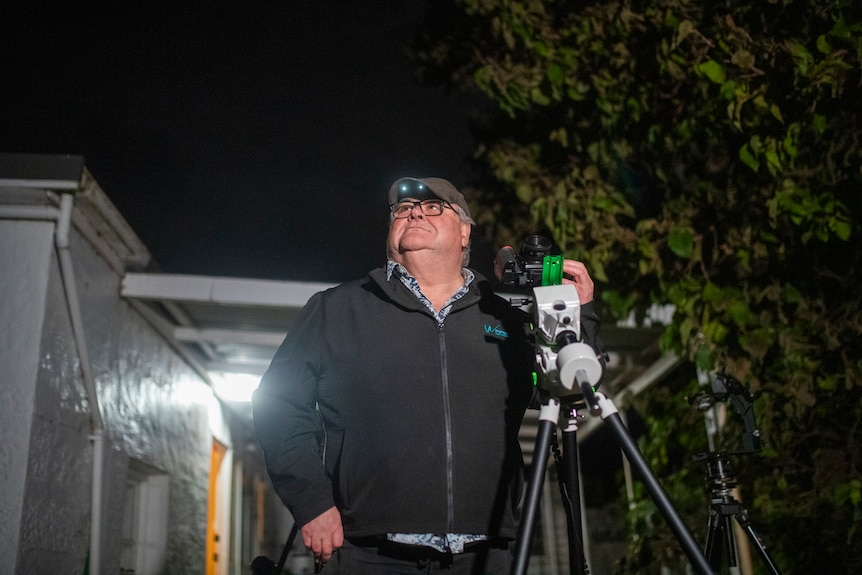A man with a head torch stands next to his camera and looks up at a dark sky.