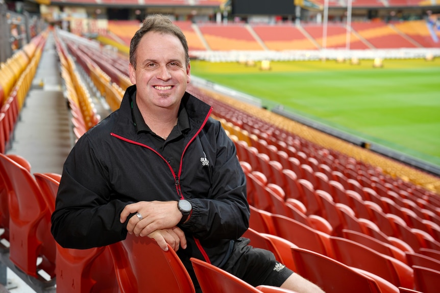 A man in a black weatherproof jacket sitting in stadium stand smiling at the camera.