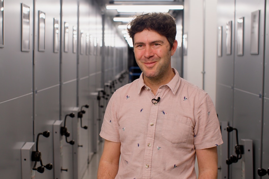 A middle aged man in a pink shirt stands in an archive room with a hall of shelves behind him.