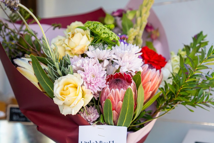 A colourful bunch of flowers sits in a white box, with burgundy paper wrapped around the bunch