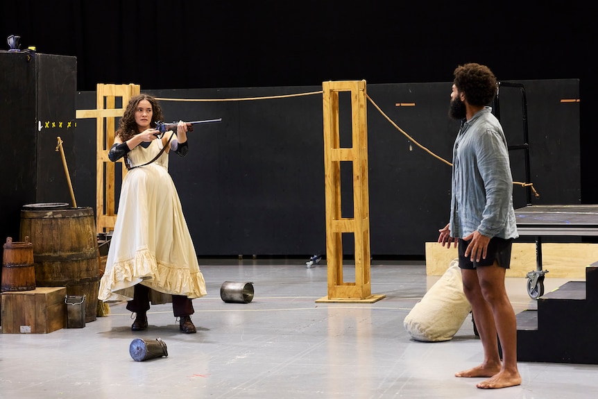 Nina Korbe dressed in a long dress over pants holds an old rifle up pointed at Marcus Corowa.