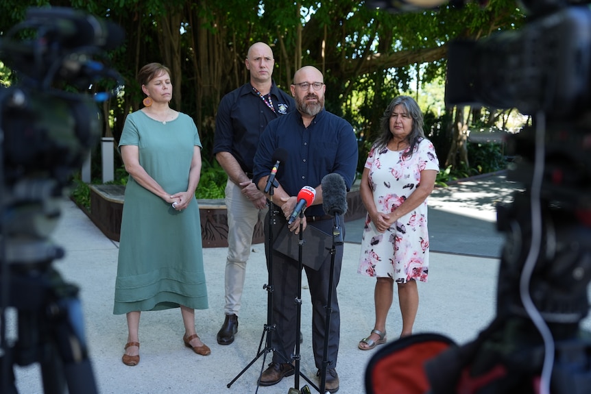 Four people stand in front of news microphones, with blurred television cameras in the foreground.