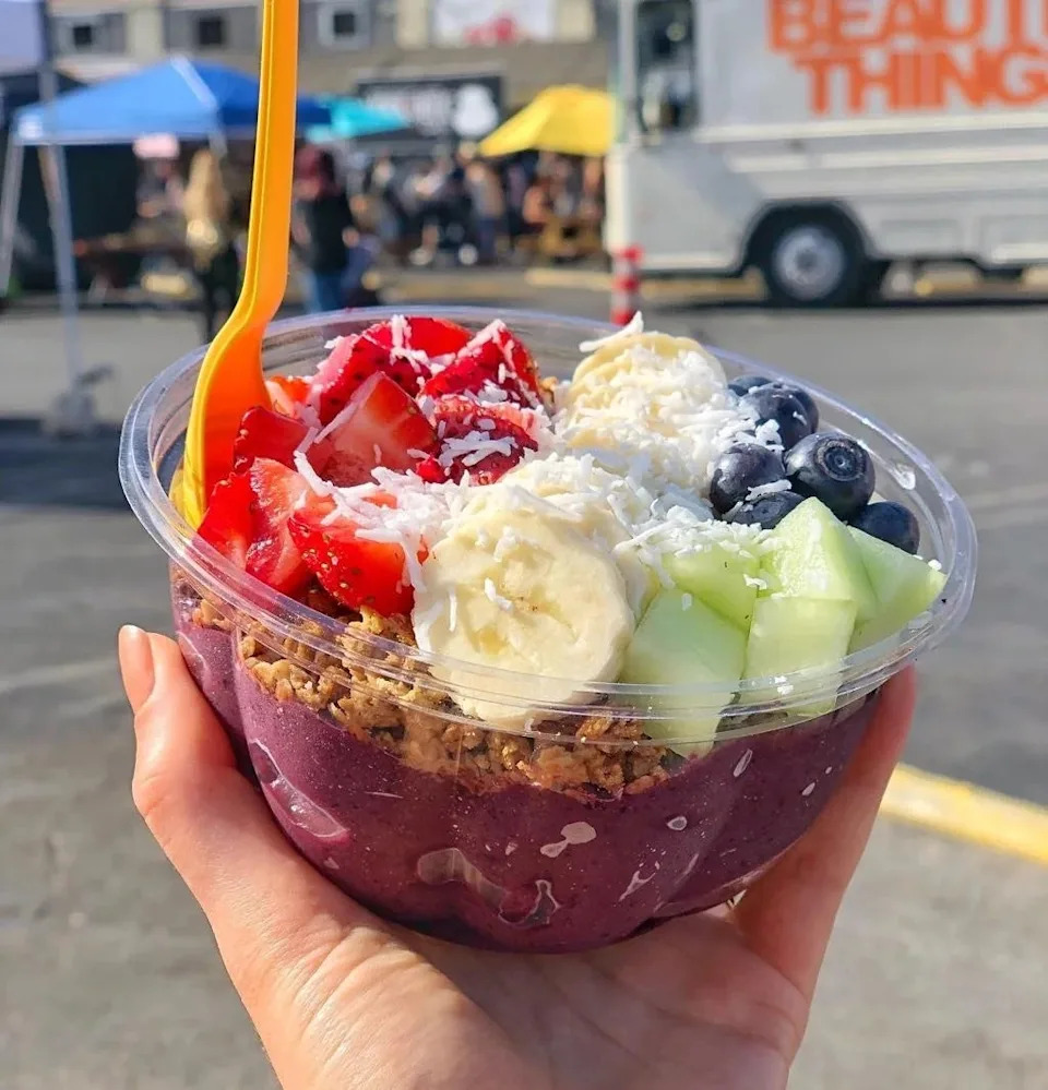 Smoothie bowl topped with sliced strawberries, bananas, blueberries, and melon in a hand. Granola and coconut flakes add texture