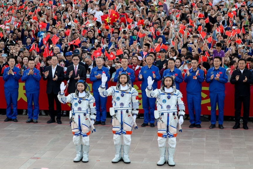 Three Chinese astronauts in white space suits smiling and waving in front of a crowd of others with Chinese flags.