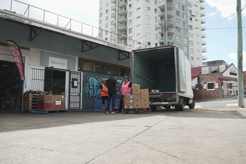 A truck parked out the front of a suburban warehouse. A man in high-vis stands next to a pallet stacked with carboard boxes.