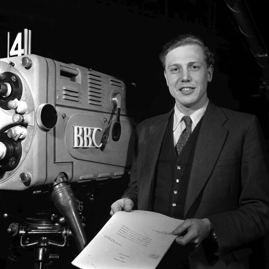 Black and white image of young David Attenborough standing in front of TV camera.