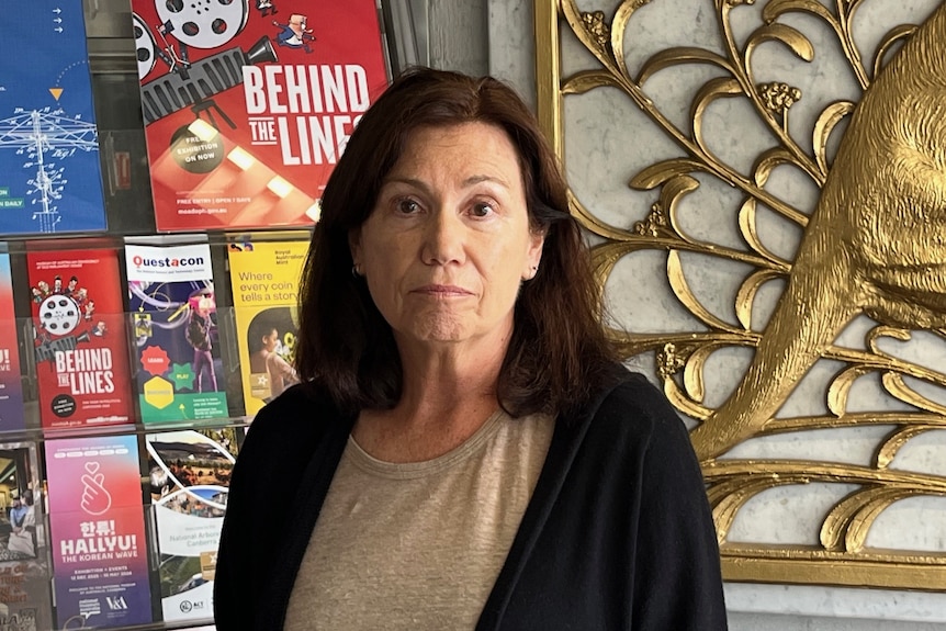 A woman with long dark hair stands indoor looking serious.