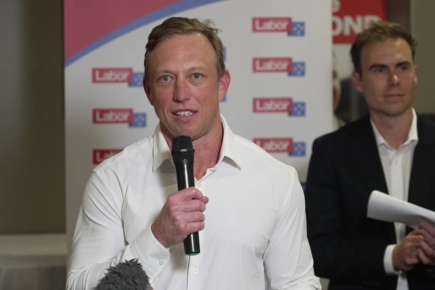 Steven Miles stands holding a microphone with Luke Richmond on the right. 