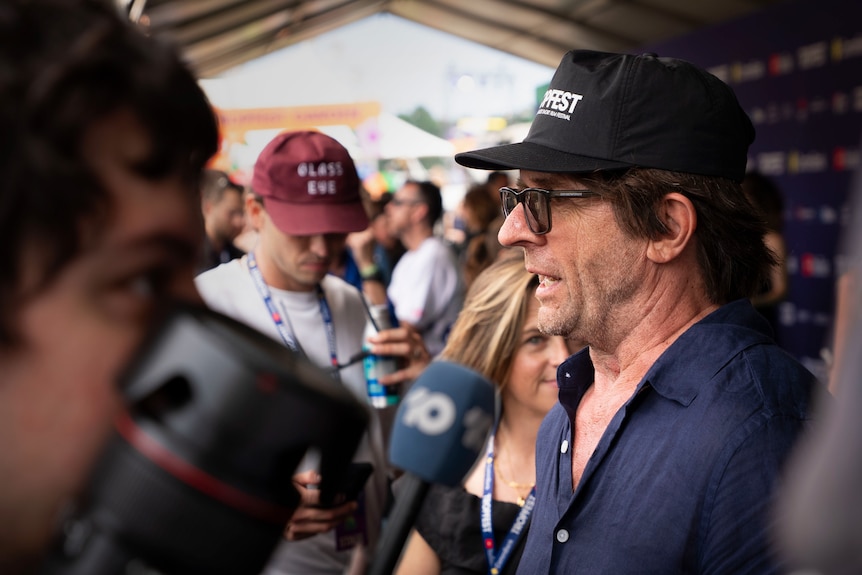 Close headshot of a man with cap and glasses side profile speaking into microphone in busy area of people and media wall