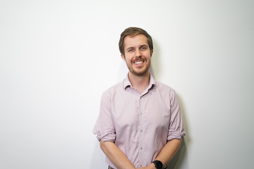 A young man with short dark blonde hair in a checked business shirt smiles at the camera.