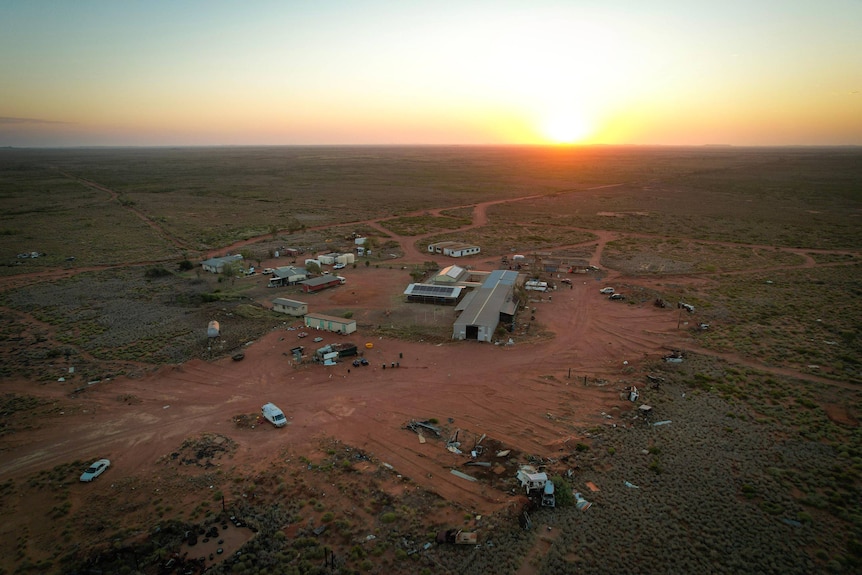 An aerial image of a haphazard compound in the red desert, with the sun setting on the horizon.