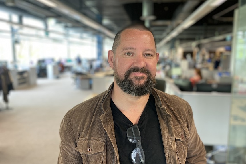 A man with short dark hair and a beard in a brown jacket over a black t-shirt with a blurred office in the background