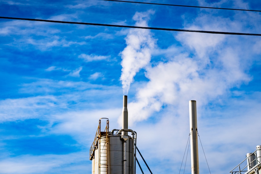 White steam comes from a steel ventilation stack near steel tanks against a blue sky with wispy white clouds.