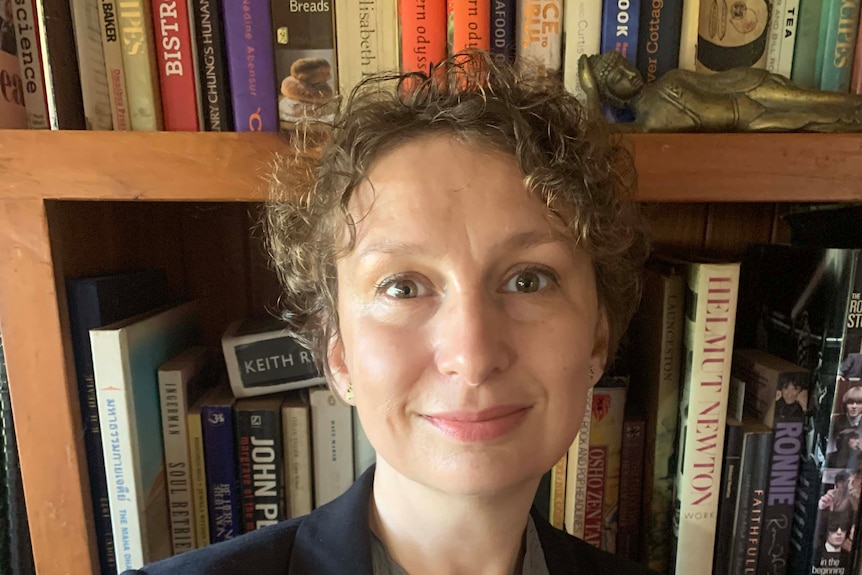 A woman wearing glasses and a suit jacket stands in front of a full book shelf.