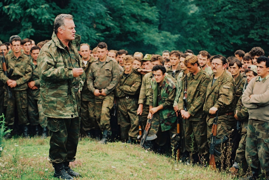 A middle-aged man with grey hair in an army jacket speaks to a large group of young soldiers in the forest.