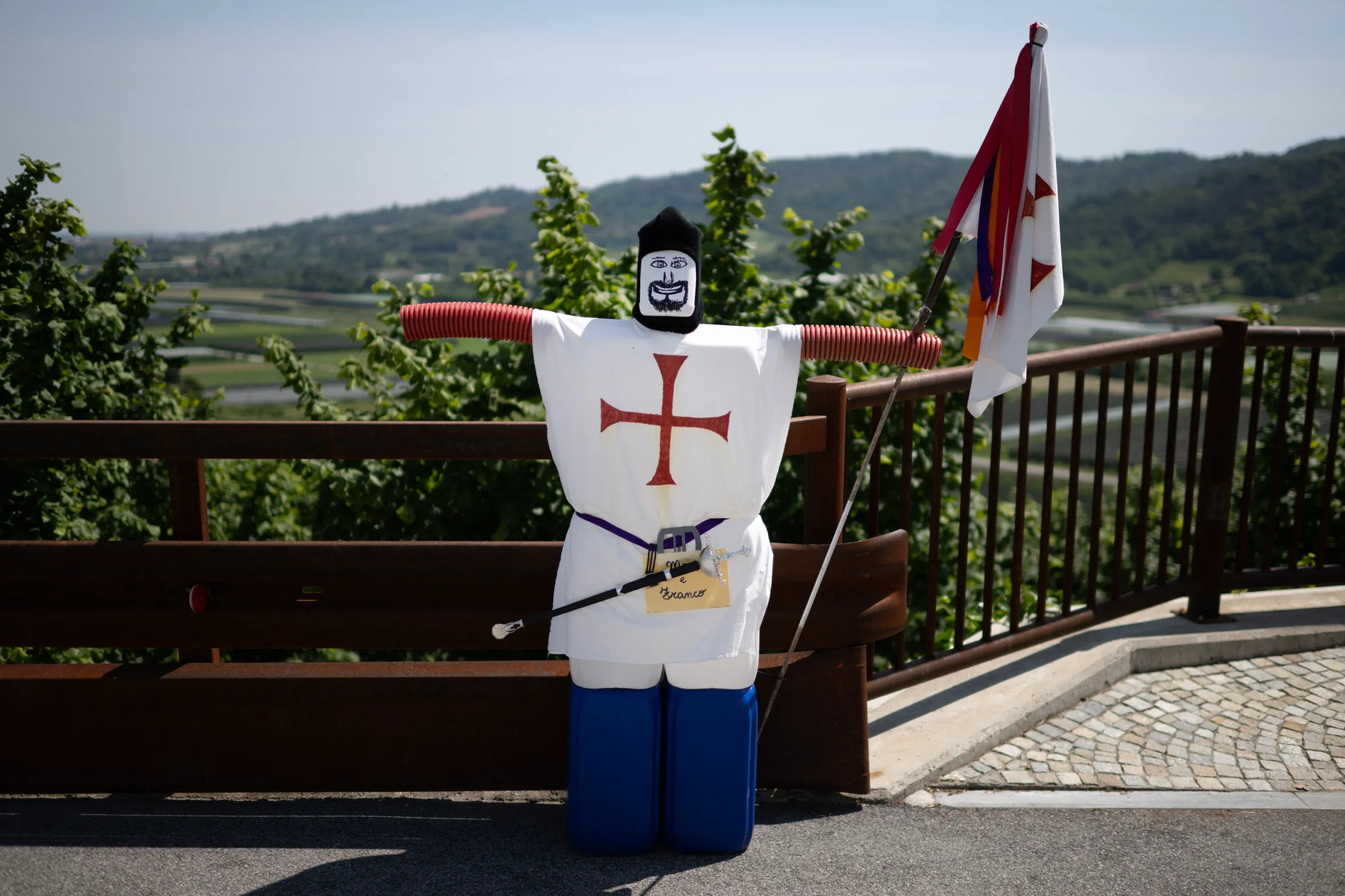 A scarecrow dressed in a white tunic with a red cross, blue pants, and a black hat with a painted face, holding a colorful flag, stands next to a wooden fence overlooking a valley.