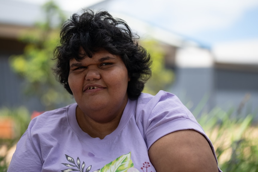 An Aboriginal girl wearing a lilac t-shirt with short black hair smiles while sitting outside a library's garden.