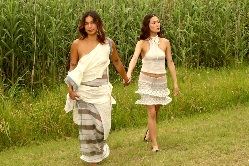 Two woman wearing white, boho outfits, hold hands and pose for photo in front of corn field