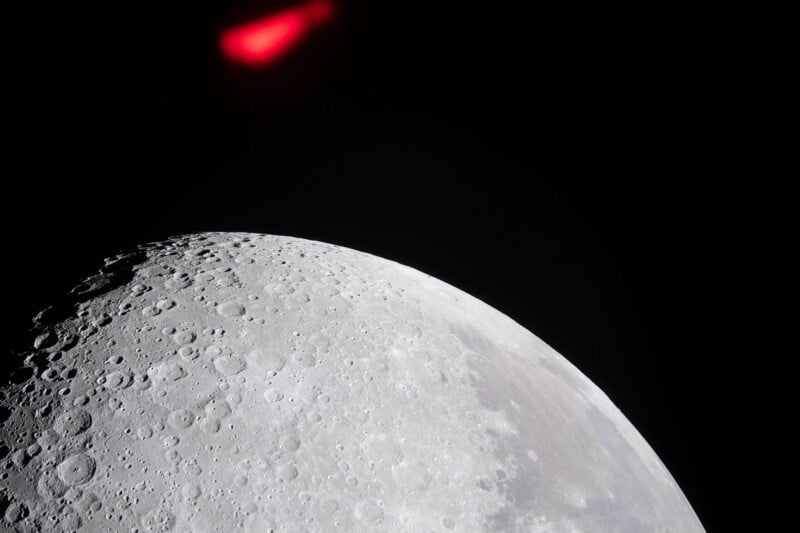 A close-up view of the moon’s cratered surface with a bright red, blurry light or object in the dark space above the moon.