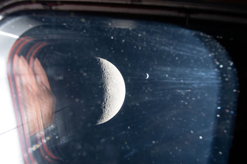 A close-up view through a spacecraft window shows the moon in the foreground and a smaller planet or moon in the distance, with a hand reflected on the window and dust particles visible.