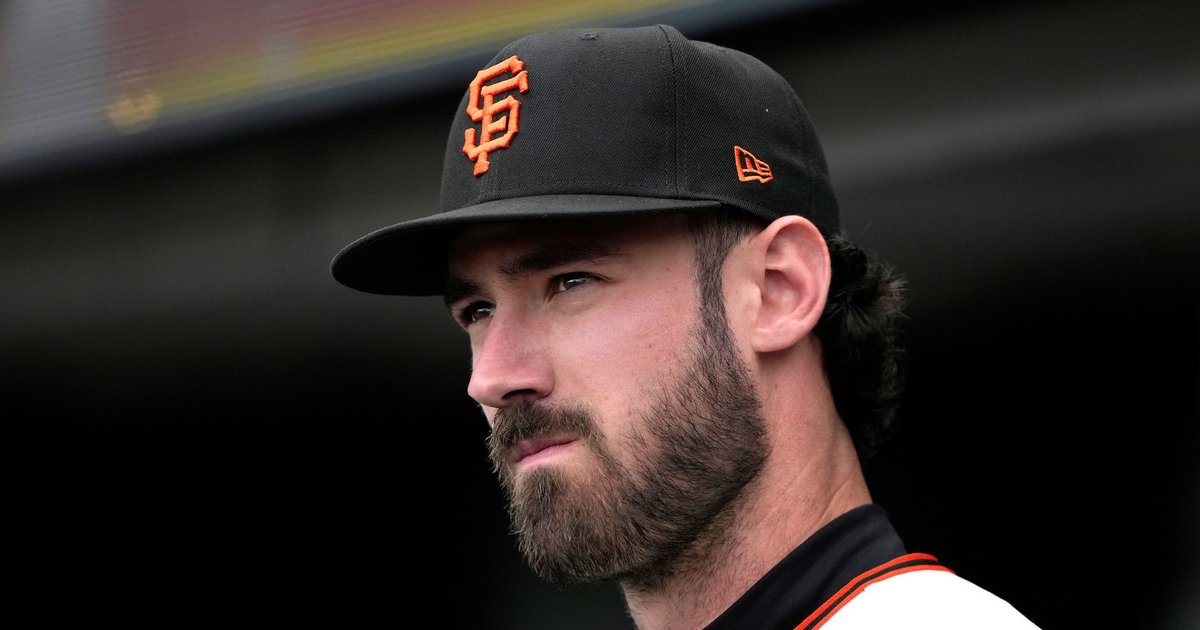 A man with a beard wearing a black San Francisco Giants baseball cap and white jersey stares thoughtfully into the distance against a dark background.