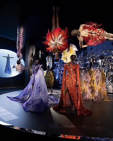 Fashion exhibit with mannequins wearing vibrant avant-garde dresses, suspended in a dark room with dramatic lighting.