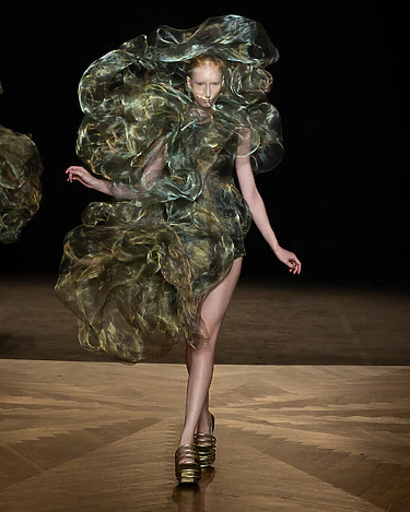 Model wearing a flowing, abstract dress with intricate textures on a runway, showcasing avant-garde fashion design.