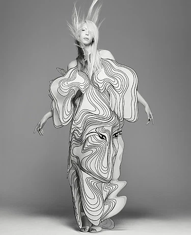 Model in intricate, flowing black-and-white patterned dress with artistic, wavy design in dynamic pose against gray background.