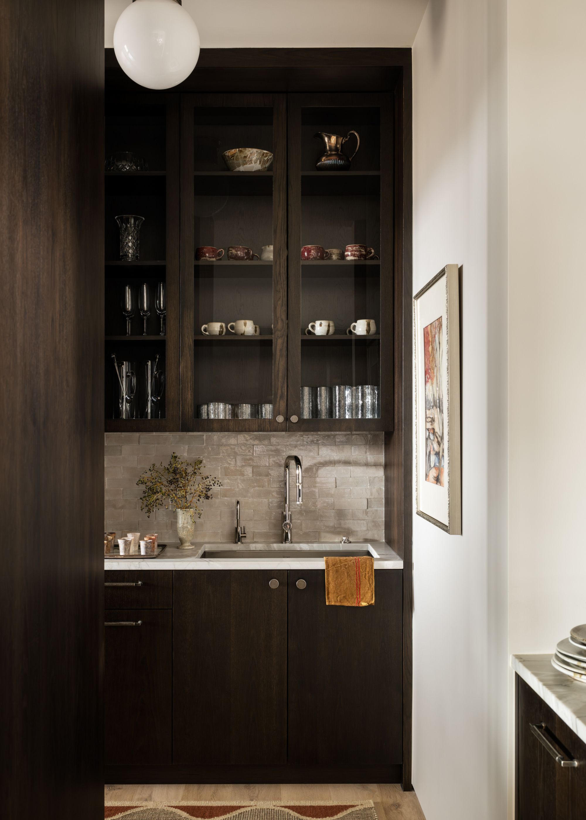 A butler's pantry in dark wood tones with glass-fronted cabinets with a rug on the floor and decorative items on the shelves